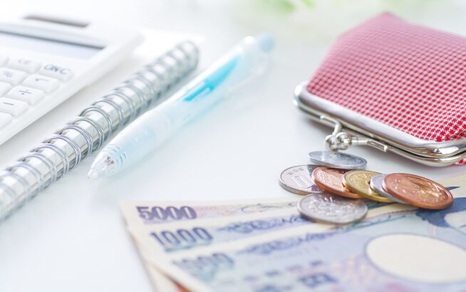 デスクに置かれた日本円の紙幣（五千円札、千円札）、硬貨、がま口財布、計算機、文房具